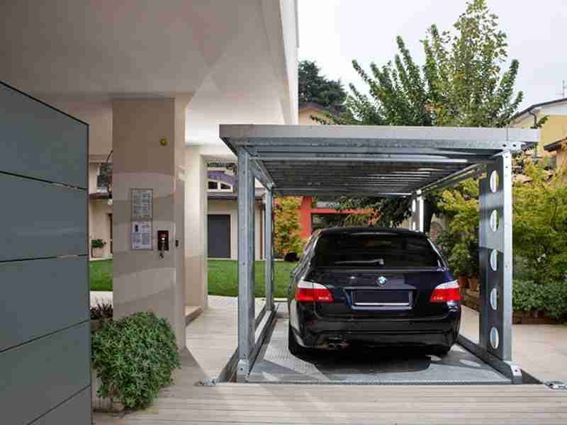 Auto nera parcheggiata sotto struttura metallica grigia con tetto, giardino e casa sullo sfondo